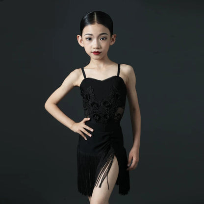 A young girl modeling a black dance dress with floral lace details and fringe, posed against a dark gray studio background 